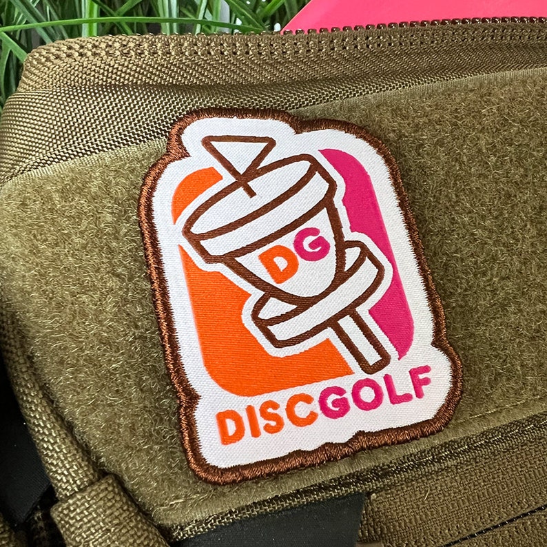 May include: A white, brown, orange, and pink embroidered patch with a disc golf basket and the text "DISCGOLF".
