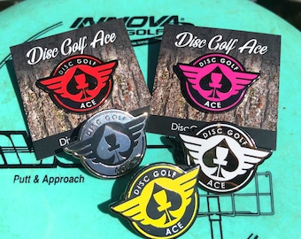 Può includere: Una collezione di spille da disc golf in vari colori, tra cui rosso, rosa, argento, bianco e giallo. Ogni spilla presenta un design a picche con le parole "Disc Golf Ace". Le spille sono esposte su un disco da disc golf verde.