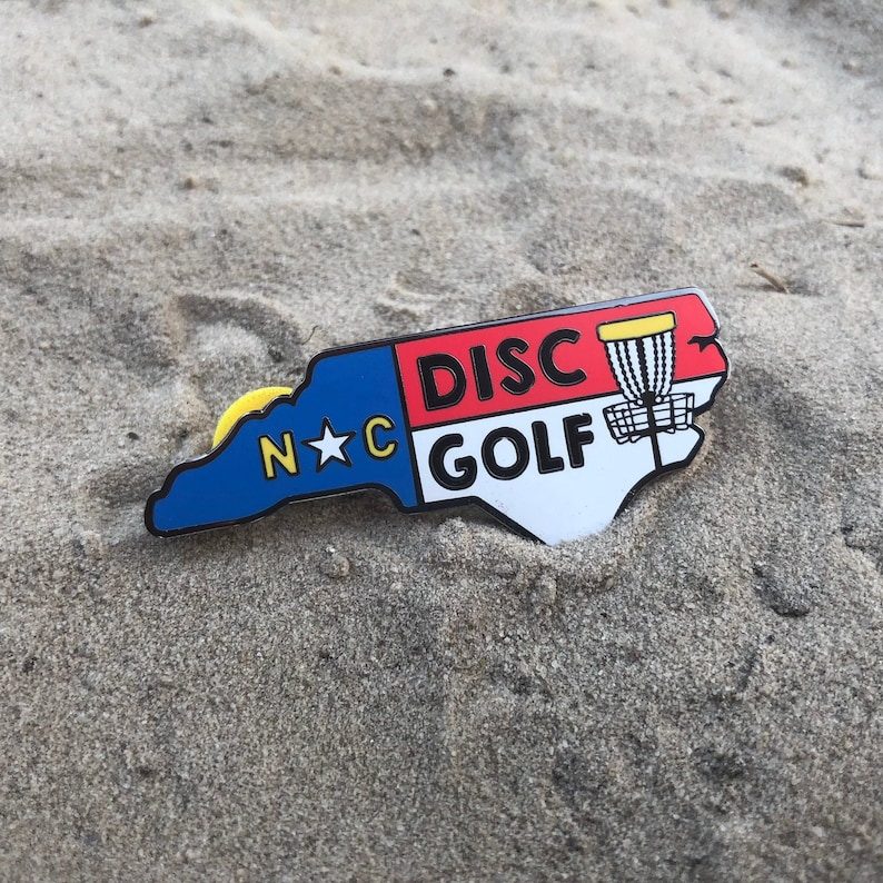 Puede incluir: Un pin con forma de Carolina del Norte azul, rojo y blanco con una estrella amarilla y el texto "N*C DISC GOLF" y una canasta de disco golf.