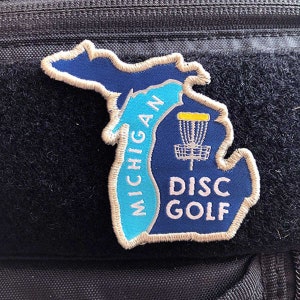 May include: A blue and white patch shaped like the state of Michigan with the words "Michigan Disc Golf" and a disc golf basket.