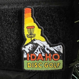 Può includere: Una toppa nera e verde con il contorno dello stato dell'Idaho. La toppa presenta un cestino da disc golf, una catena montuosa e il testo "IDAHO DISC GOLF".