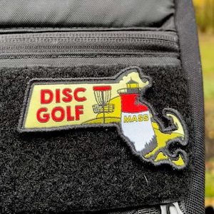May include: A yellow and red patch with the text "DISC GOLF" and a silhouette of the state of Massachusetts with a lighthouse and the text "MASS".