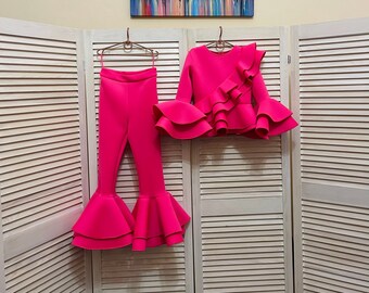 Puede incluir: Un conjunto de dos piezas de color rosa brillante con una blusa con volantes y pantalones acampanados. La blusa tiene un solo volante en un hombro y varios volantes en la parte inferior. Los pantalones tienen una pierna ancha y dos capas de volantes en la parte inferior.