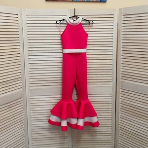 Peut inclure: Une combinaison rose et blanche avec un haut halter et un ourlet à volants. La combinaison est suspendue à un cintre sur un fond blanc à lattes.
