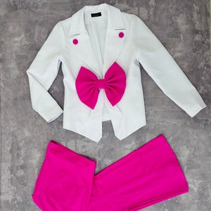 Puede incluir: Un blazer blanco con un gran lazo fucsia y pantalones fucsia a juego. El blazer tiene dos botones fucsia y un corte entallado. Los pantalones son de estilo recto.
