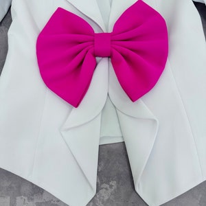 May include: White blazer with a large fuchsia bow detail. The blazer has a classic design with a single button closure and lapels. The bow is a prominent feature, adding a bold contrast to the clean lines of the jacket.