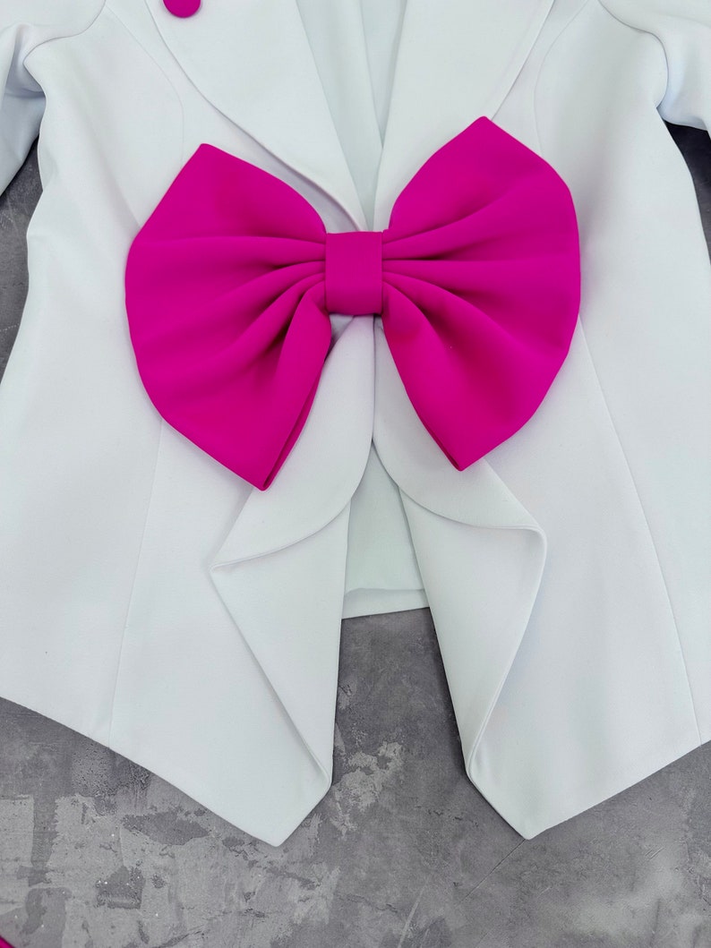 May include: White blazer with a large fuchsia bow detail. The blazer has a classic design with a single button closure and lapels. The bow is a prominent feature, adding a bold contrast to the clean lines of the jacket.