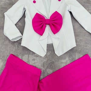 May include: A white blazer with a large fuchsia bow detail and matching fuchsia pants. The blazer has two fuchsia buttons and a notched lapel. The pants are wide-legged and made of a smooth fabric. The outfit is laid out on a gray surface.