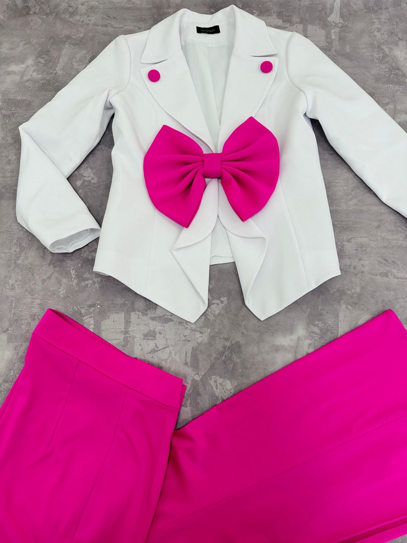 May include: A white blazer with a large fuchsia bow detail and matching fuchsia pants. The blazer has two fuchsia buttons and a notched lapel. The pants are wide-legged and made of a smooth fabric. The outfit is laid out on a gray surface.