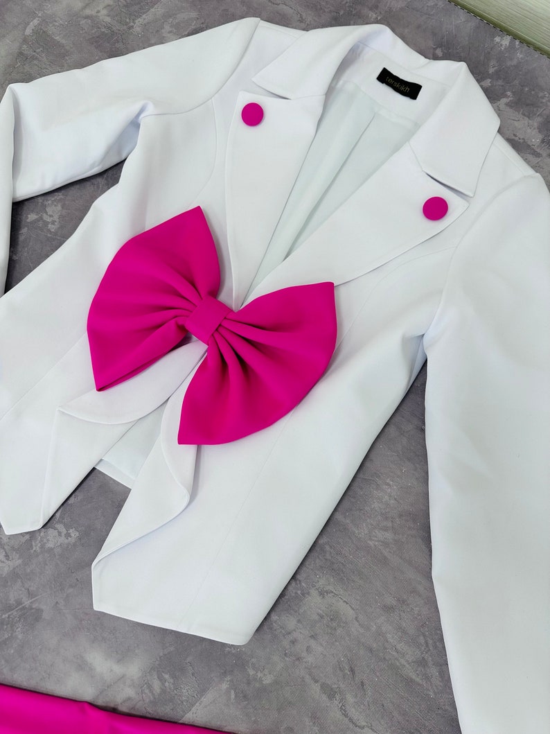May include: A white blazer with a large, bright pink bow and matching pink buttons. The blazer has a classic tailored design, with a notched lapel and long sleeves. The bow is a prominent feature, adding a bold pop of color.