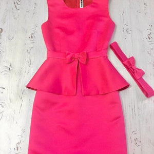 May include: A bright pink sleeveless dress with a bow at the waist and a matching pink headband. The dress has a fitted bodice and a full skirt.
