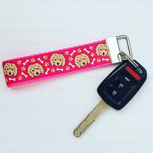 May include: Pink key chain with a pink and white ribbon featuring a repeating pattern of brown dogs and white bones. The key chain is attached to a black and silver car key.