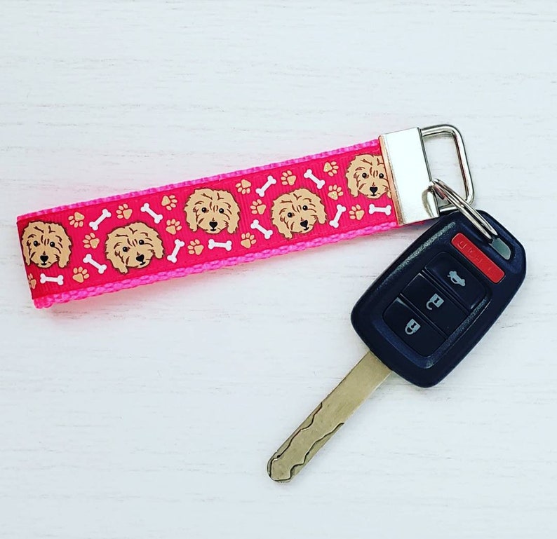 May include: Pink key chain with a pink and white ribbon featuring a repeating pattern of brown dogs and white bones. The key chain is attached to a black and silver car key.