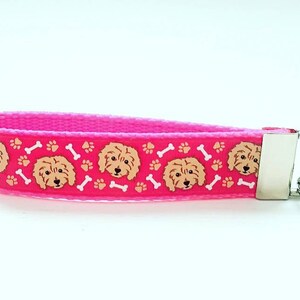 May include: Pink key chain with a repeating pattern of brown doodle dog faces, white paw prints, and white bone shapes.