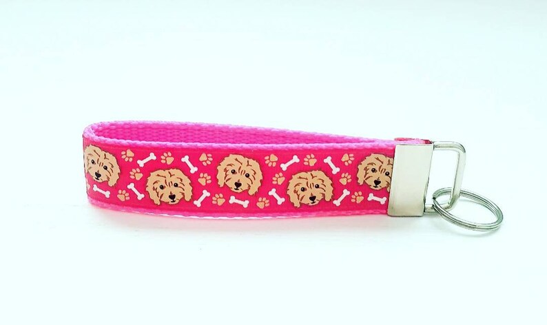May include: Pink key chain with a repeating pattern of brown doodle dog faces, white paw prints, and white bone shapes.