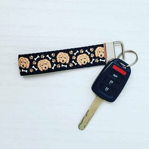 May include: Black and white key chain with a repeating pattern of brown dog faces and white bone shapes. The key chain is attached to a black car key with a red button.