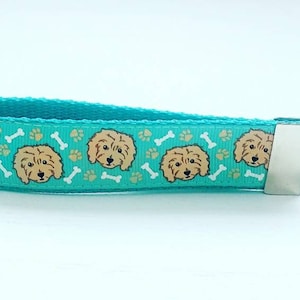 May include: A turquoise blue key chain with a repeating pattern of brown doodle dog faces and white bone shapes. The key chain has a silver metal key ring.