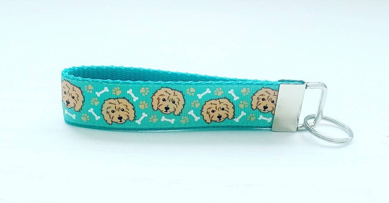 May include: A turquoise blue key chain with a repeating pattern of brown doodle dog faces and white bone shapes. The key chain has a silver metal key ring.