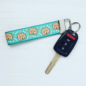 May include: A teal blue key chain with a repeating pattern of brown doodle dogs and white bone shapes. The key chain is attached to a black car key with four buttons.
