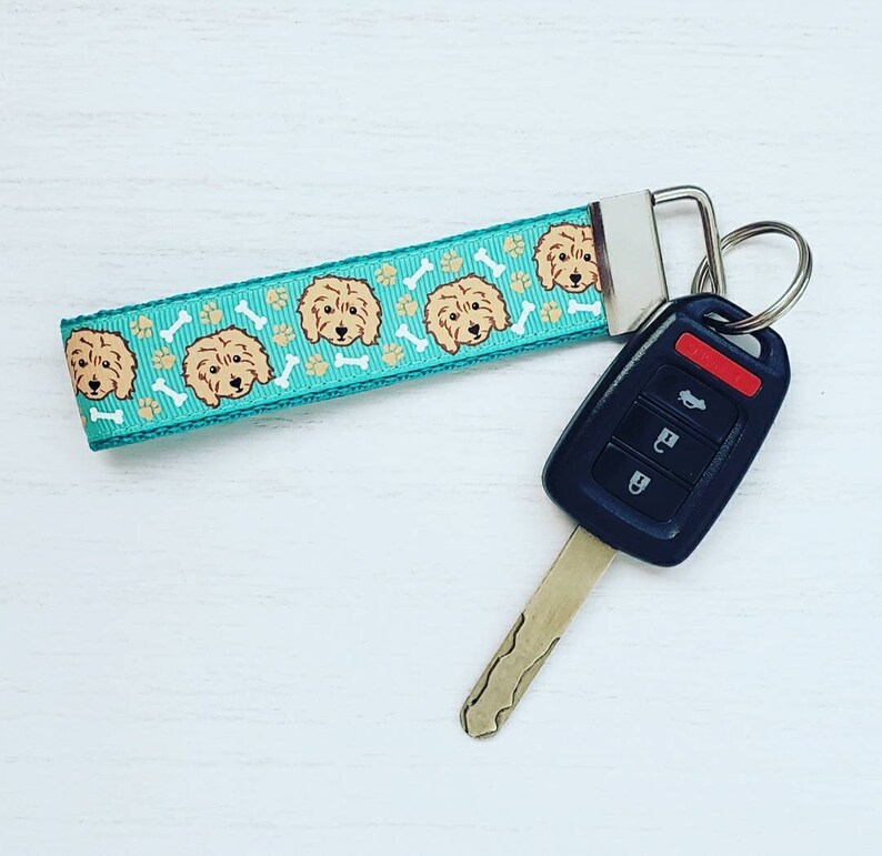May include: A teal blue key chain with a repeating pattern of brown doodle dogs and white bone shapes. The key chain is attached to a black car key with four buttons.