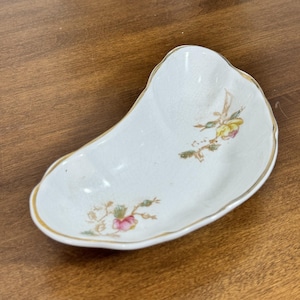 May include: White ceramic dish with a gold rim and floral accents. The dish has a unique, elongated shape and features delicate pink and yellow flowers with green leaves. A vintage-style decorative piece.