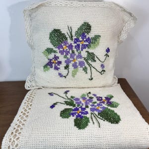 May include: A square off-white crochet pillow and a matching folded throw blanket, both featuring a cross-stitch pattern of purple and violet flowers with green leaves. The edges of the pillow and the blanket are finished with a decorative crochet border.