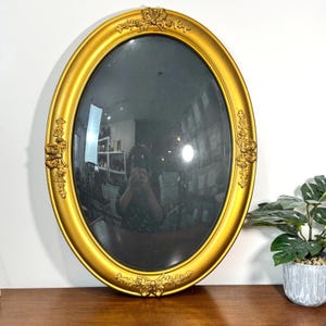 Può includere: Specchio ovale dorato con dettagli decorativi. La cornice ha una finitura lucida e accenti floreali decorativi. La superficie dello specchio riflette l'ambiente circostante.