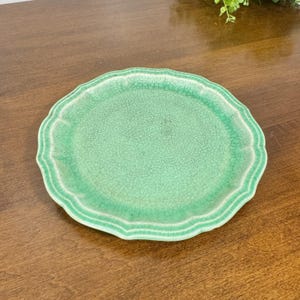 May include: A round ceramic plate featuring a vibrant mint green glaze with a prominent crackle finish. The plate has a scalloped, wavy edge design that creates a decorative border around the central surface, showcasing a detailed network of fine lines.