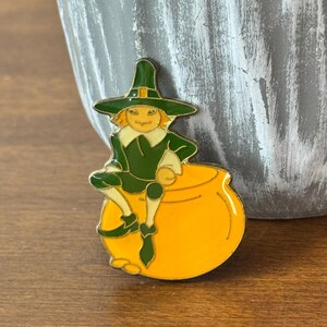 Peut inclure: Une épingle colorée en émail représentant un personnage de dessin animé de lutin vêtu de vert et portant un chapeau pointu, assis sur un pot d'or jaune. L'épingle a une bordure dorée et est posée sur un fond neutre.