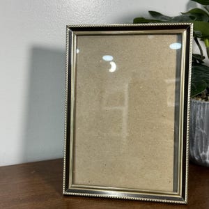 May include: A rectangular picture frame with a gold-colored beaded border and a dark brown inner frame. The frame has a glass front and a brown backing. The frame is standing upright on a wooden surface.
