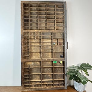 May include: A vintage wooden printer's tray with many small compartments. The tray is made of aged wood with a natural finish, showing signs of wear. A few small objects are placed in some of the compartments. The tray is displayed on a wooden surface.