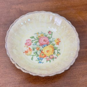 May include: A small, round, yellow bowl with a scalloped edge and a floral cross-stitch design in the center. The flowers are pink, yellow, orange, and blue. The rim has a gold trim.
