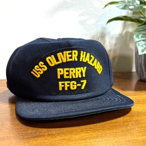 May include: A navy blue baseball cap with a flat brim. The cap features a yellow embroidered patch with the text "USS OLIVER HAZARD PERRY FFG-7". The cap is sitting on a wooden surface.