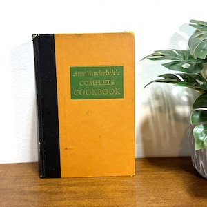 May include: A vintage cookbook titled "Amy Vanderbilt's COMPLETE COOKBOOK." The book has a mustard yellow cover with a green rectangular label. The spine is black. The book is standing upright on a wooden surface.