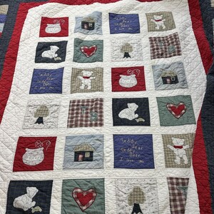 May include: A patchwork quilt with a white background and red border. The quilt features squares with teddy bears, hearts, houses, and trees. Some squares have the text "Teddy Bear I love you".