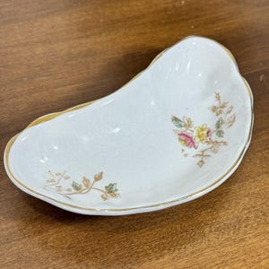 May include: White ceramic dish with a gold rim and floral design. The dish has a kidney bean shape and features a delicate pattern of pink, yellow, and green flowers with brown stems. The dish is likely vintage.