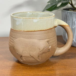 Könnte beinhalten: Handgefertigte Keramiktasse in Beige- und Cremetönen. Die Tasse zeigt ein erhabenes Relief eines Schweins an der Seite. Henkel und Rand sind cremefarben, der Körper ist beige. Ideal für Kaffee oder Tee.