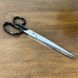 May include: A pair of scissors with black handles and silver blades. The scissors are lying on a wooden surface. The blades are slightly open, ready for use.