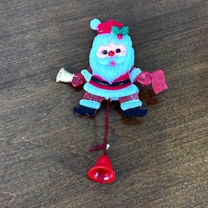 May include: A Santa Claus brooch featuring a red hat with green holly, a white beard, a red suit with black trim, and black boots. The figure holds a gold bell in one hand and a red scroll in the other, with a red string hanging down to a red bell.