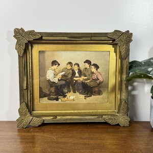 May include: A framed print depicting five young boys gathered together, with one reading a paper. The frame is gold-colored with leaf-shaped corner accents. The print has a warm, vintage aesthetic.