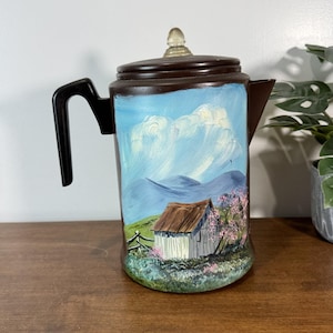 May include: A brown vintage coffee pot with a black handle and a clear knob on the lid. The pot is painted with a landscape scene featuring a white barn, mountains, and a blue sky with clouds.