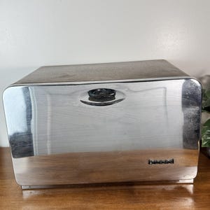 Op de afbeelding: Een vintage rechthoekige broodtrommel met een glanzende, zilverkleurige metalen buitenkant. De doos heeft een gebogen voorkant en een kleine, ovale handgreep aan de bovenkant. Het woord "bread" staat in zwarte letters op de voorkant.