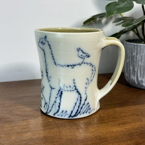 May include: A cream-colored ceramic mug featuring a hand-drawn dark blue illustration of a llama with a small bird perched on its back. The mug has a smooth, glossy finish, a sturdy handle on the right side, and a slightly flared rim at the top.