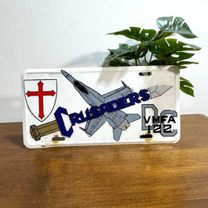 May include: A white license plate featuring a Crusader theme. The plate has a red cross and a stylized jet, with the word "CRUSADERS" and "DC VMFA 122" in blue and gray. The plate has a distressed look.