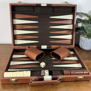 Puede incluir: Juego de backgammon de cuero marrón abierto, que revela el tablero de juego. El tablero tiene triángulos marrones y crema. Incluye dados, cubiletes y fichas. El juego se muestra abierto, listo para jugar.