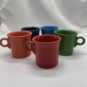 Op de afbeelding: Vijf keramische mokken in verschillende kleuren: rood, oranje, groen, blauw en donkerblauw. Elke mok heeft een enkele handgreep met een ringvorm.