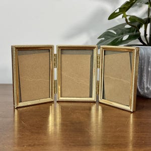 May include: A gold-colored, tri-fold picture frame with a rope-like border. The frame is open, revealing three sections with a cardboard backing. The frame is on a wooden surface, with a plant in the background.