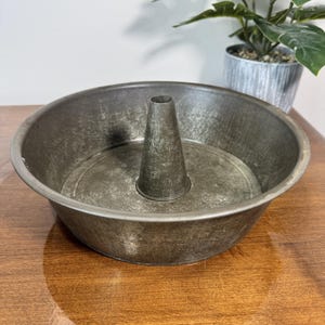 Puede incluir: Un molde para bizcocho bundt redondo de metal vintage con un cono central. El molde tiene un aspecto envejecido y desgastado con un acabado metálico oscuro. Se encuentra sobre una superficie de madera, lo que sugiere un tema de cocina o repostería. Diámetro aproximado de 25 cm.