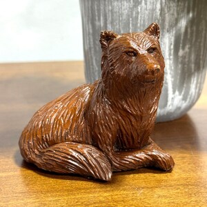 May include: A detailed, brown wooden bear figurine in a seated position. The bear has textured fur and is positioned on a wooden surface. The figurine is a decorative item, possibly for a shelf or desk.