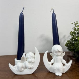 May include: Two white ceramic candle holders, each featuring a small angel figure resting on a crescent moon. Each holder supports a tall, dark navy blue taper candle. The figures are smooth, glossy, and sculpted with detailed wings.